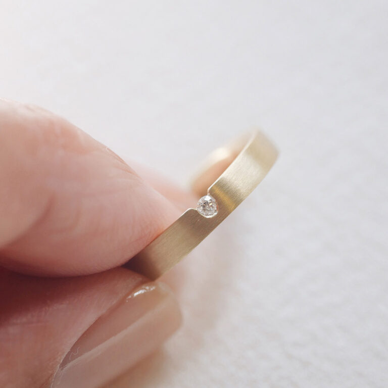 mão segurando aliança de casamento feita a mão em ouro 18k e diamante central de 3 pontos minimalista sobre fundo branco