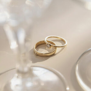 Par de alianças minimalistas feitas a mão em ouro 18k com diamante para casamentos ao lado de taça de vidro