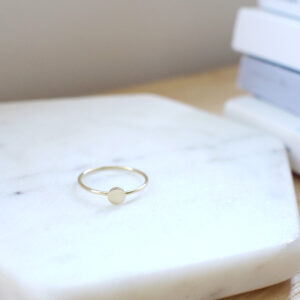 Anel minimalista de aro fino feito a mão em ouro 18k com disco na frente em cima de superfície de mármore
