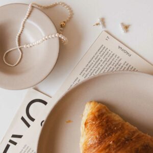 Joias minimalistas com pérolas naturais de tamanhos diferentes sobre uma mesa com croissant, pratos e jornal