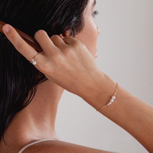 Mulher de cabelos pretos usando pulseira, anel e brincos de ouro 18k minimalistas feitos a mão com pérolas naturais penteando o cabelo