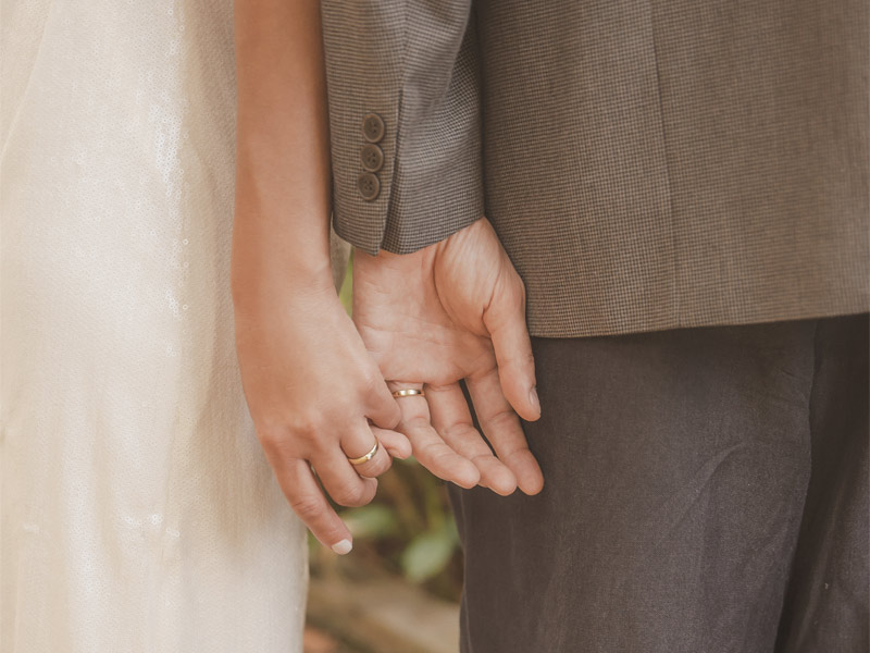 Mulher de vestido de noiva e homem de terno, com alianças nos dedos e mãos dadas levemente.