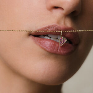 foto com foco na boca de uma mulher onde na frente está pendurado um colar de ouro 18k e um pingente de coração cravejado com 14 rubis